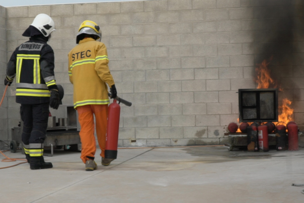 Dos bomberos equipados con trajes de protección completos practican técnicas de rescate y control de incendios durante un entrenamiento. Uno de ellos sostiene una extintor mientras el otro lo respalda, asegurándose de la correcta posición y manejo del equipo. En el fondo, se observa un fuego controlado utilizado para el simulacro, con humo denso que añade realismo a la práctica. La escena destaca la importancia de la coordinación y las habilidades adquiridas en la formación para enfrentar emergencias de manera segura y efectiva.