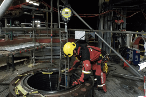 Un bombero visto desde arriba asiste a su compañero que está saliendo de un espacio confinado, sosteniendo firmemente una cuerda de seguridad conectada al arnés. Lleva equipo de protección completo y un casco con linterna frontal. En el borde de la entrada estrecha, se aprecian herramientas de rescate y un detector de gases cercano, destacando la supervisión constante y las medidas preventivas necesarias para la evaluación de riesgos.