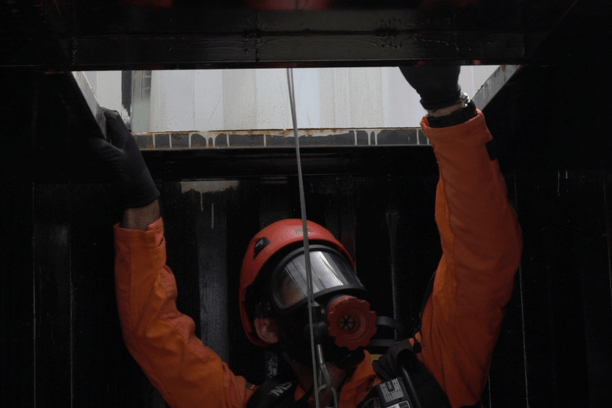 Un bombero equipado con traje de protección, máscara de respiración y arnés de seguridad emerge cuidadosamente de un espacio confinado, como una abertura estrecha de un tanque o conducto industrial. Sus movimientos son guiados por un compañero que lo asiste desde el exterior, asegurando las cuerdas de seguridad. 